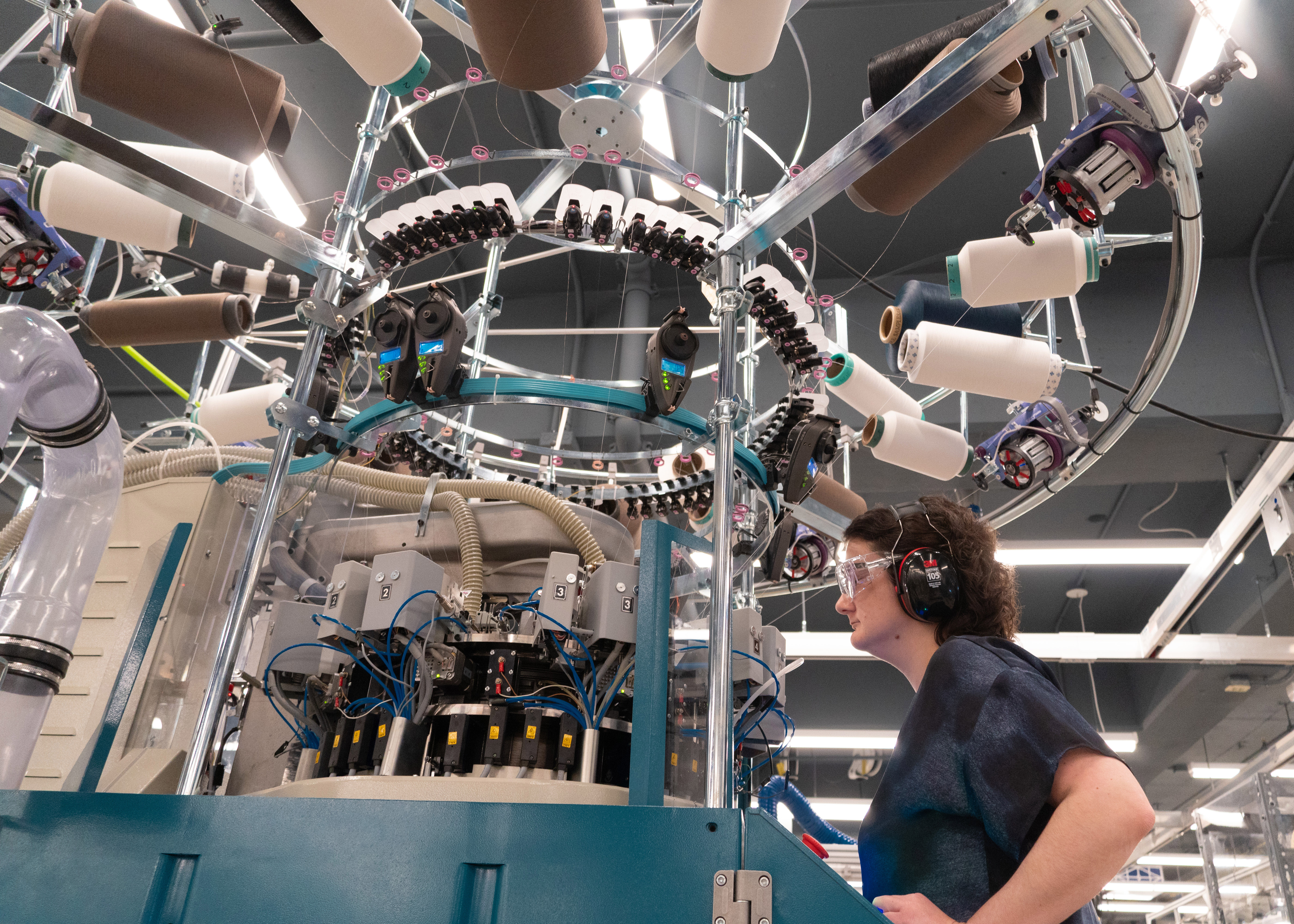 A person wearing industrial ear and eye protection operating a large circular weft knitting machine